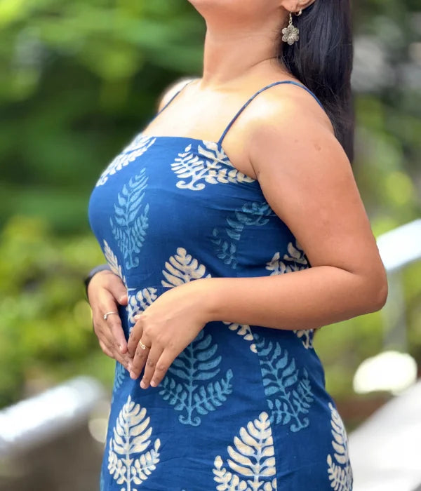 Woman in an indigo blue cotton bodycon dress with white hand-block leaf prints, strappy neckline, and a side slit, standing outdoors in a lush green setting.