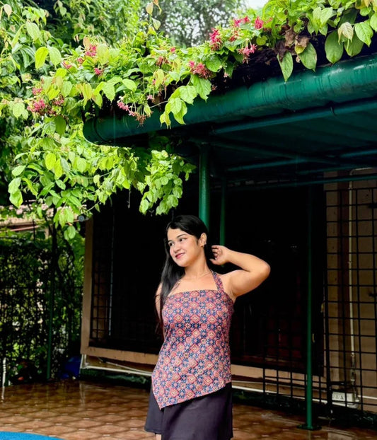 Woman posing outdoors in a stylish backless halter neck top with an asymmetrical hem, tie-up back, and shorts, surrounded by lush green plants and a poolside patio. Perfect summer fashion vibe.