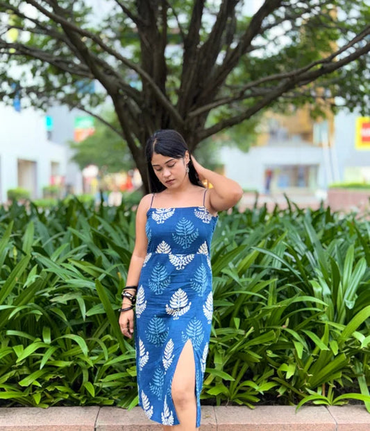 Woman in an indigo blue cotton bodycon dress with white hand-block leaf prints, strappy neckline, and a side slit, standing outdoors in a lush green setting.