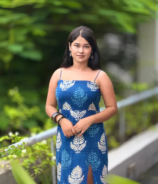 Woman in an indigo blue cotton bodycon dress with white hand-block leaf prints, strappy neckline, and a side slit, standing outdoors in a lush green setting.
