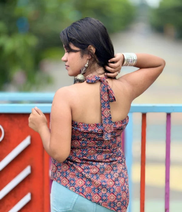 Woman posing outdoors in a stylish backless halter neck top with an asymmetrical hem, tie-up back, and shorts, surrounded by lush green plants and a poolside patio. Perfect summer fashion vibe.