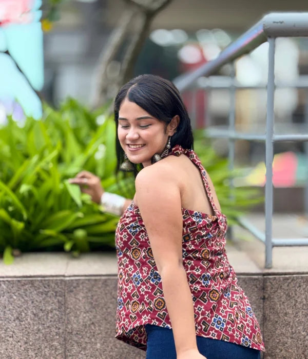 Woman wearing a bold backless halter neck top with asymmetrical hem and tie-up back, styled with blue jeans in a garden setting. Modern and breezy vibe.